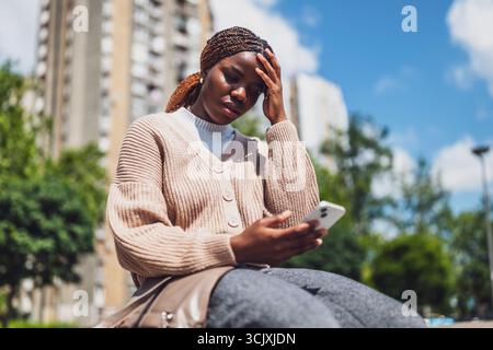 Young black woman is distraught while using smartphone for online messaging. Stock Photo