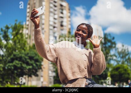 Young happy Black woman is using smartphone. She is laughing and waving while having a video call. Stock Photo