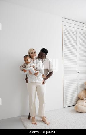 Loving biracial family holding their daughter and embracing against a white wall. Concept of interracial family. Stock Photo
