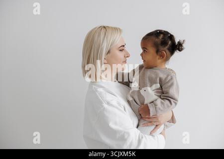 Tender side-profile portrait of a loving mother holding her biracial daughter, with both looking at each other. Concept of interracial family. Stock Photo
