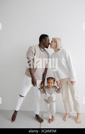 Loving biracial family holding hands their daughter sharing a tender moment of kiss against a white wall. Concept of interracial family. Stock Photo