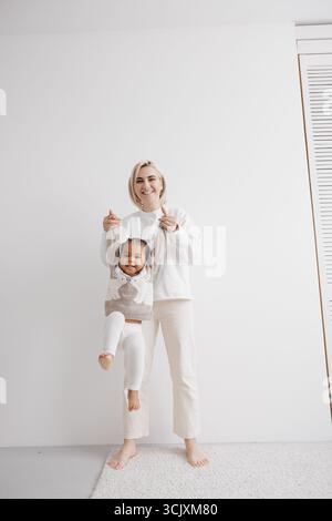 Happy mother plays and holds her biracial toddler daughter, who is smiling cheerfully. Concept of interracial family. Stock Photo