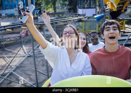 Young diverse friends are having fun on a roller coaster, taking selfies and laughing, with colorful amusement park rides in the background creating a Stock Photo