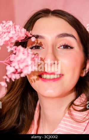 Female with long brown hair smiles softly while holding delicate pink flowers against a pastel pink backdrop, creating a peaceful and beautiful scene Stock Photo