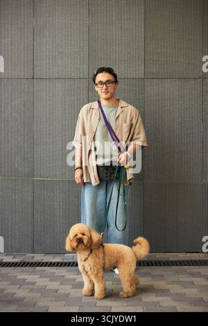 Portrait of young adult Asian man standing and holding dog leash, looking at camera with curly haired poodle standing in front, urban background with tiled wall Stock Photo