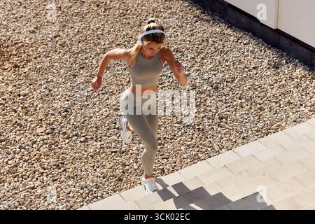 A fit young woman wearing a gray athletic outfit and running up stairs outdoors. She is energetically exercising under bright sunlight, showcasing her Stock Photo