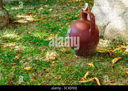 An old broken clay jug standing on the ground near a stone, surrounded by grass and fallen autumn leaves, symbolizing tradition, decay and rustic coun Stock Photo