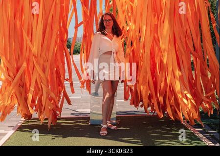 Stylish woman in a white outfit standing confidently in an outdoor installation with long, flowing orange ribbons in sunlight. Stock Photo