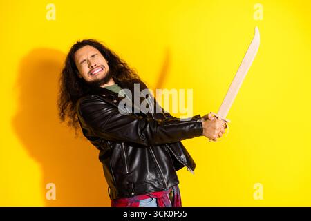 Bearded Young Man in Leather Jacket Adopting a Dramatic Pose With Wooden Sword Against Bright Yellow Background Stock Photo