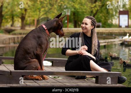 A woman with braids is seated outdoors on a wooden deck by a pond, sharing a moment with her large Doberman dog. The background features tranquil gree Stock Photo