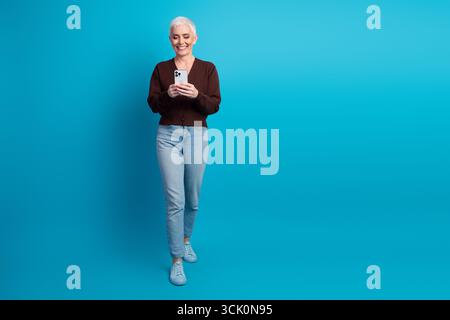 Smiling senior woman using smartphone while walking confidently against vibrant blue background in casual yet stylish clothing Stock Photo