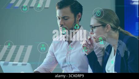 Reviewing energy data on laptop at office, two professionals wearing blazer with holographic icons Stock Photo