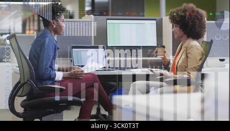 Reviewing data while two women holding coffee cups at office desk, dual monitors displaying graphs Stock Photo