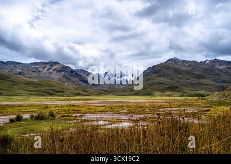 A marshy lake reflecting the dramatic cloudy sky is surrounded by rolling hills and snow-capped peaks on the Pastoruri Glacier trek in Huaraz, Peru. T Stock Photo