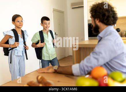 Two children with backpacks are talking to their father in modern kitchen. Excitement fills the air as they discuss their day ahead. Stock Photo