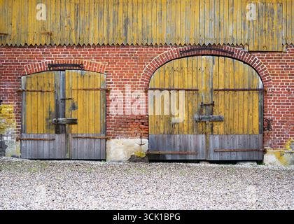 Two large rustic barn doors, weathered yellow wood framed by red brick arches, stand side by side. Stock Photo