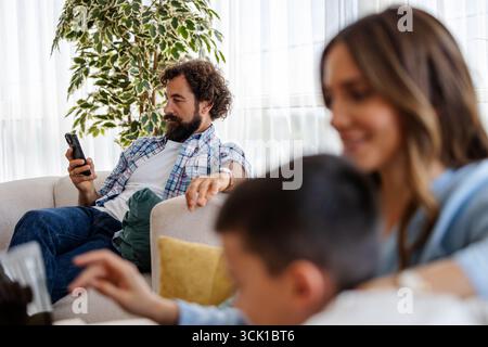 Mother enjoys relaxed moment with her son on sofa while father looks at phone with focus in their cozy living room Stock Photo