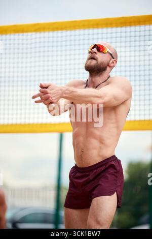 A muscular man passionately plays volleyball on an outdoor court on a sunny day. Clad in athletic gear, he demonstrates swift action and determination Stock Photo