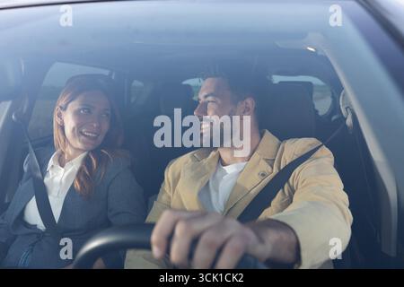 Man and woman in formal attire looking at each other and smiling while sitting in a modern car. Partners going on a business trip Stock Photo