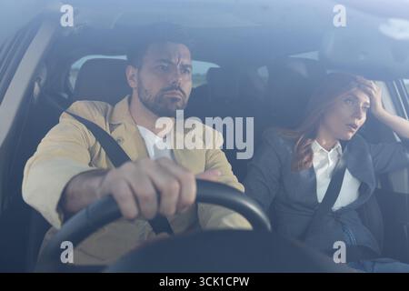 Man in drivers seat with annoyed expression gripping steering wheel, while upset woman in passenger seat holds her head. Tense atmosphere after an arg Stock Photo