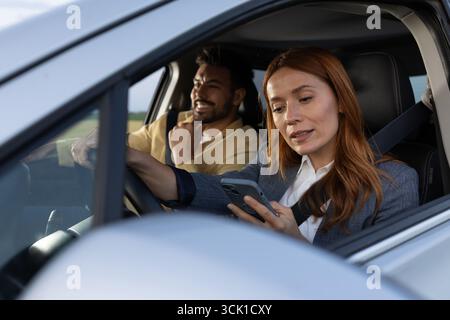 Two coworkers seated in a modern car. Woman in driver's seat distractedly looking at phone while the man is smiling Stock Photo