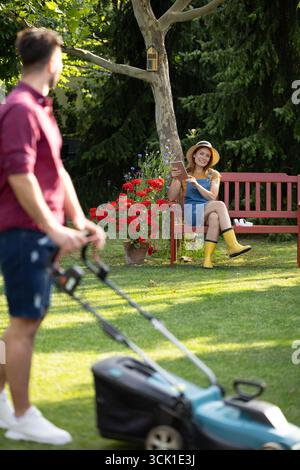 Workers in a park. Focus on a smiling female gardener sitting on a bench and taking a break from work, the man is mowing lawn and looking back at woma Stock Photo