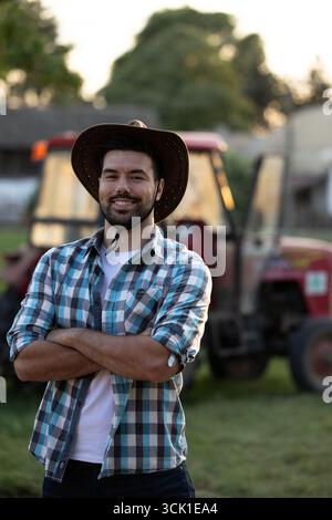 Handsome farmer standing with his arms crossed with confidence in front of tractor on ranch, cowboy hat on his head Stock Photo