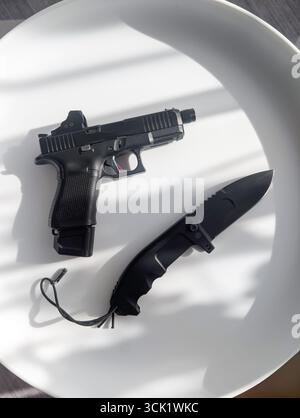 A modern semi-automatic pistol with an red dot sight and a black tactical folding knife displayed on a bright white surface. Stock Photo