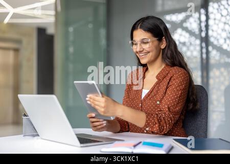 Happy businesswoman sitting at her modern office desk, using a digital tablet and laptop for work. Engaging with technology while smiling, showcasing professionalism and success Stock Photo