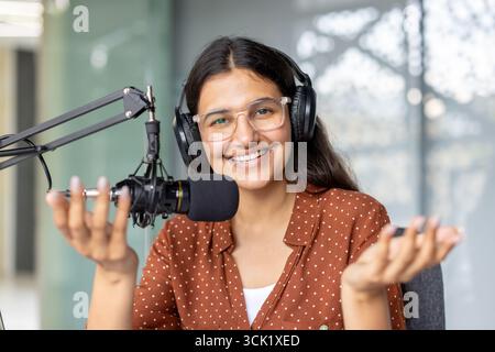 Young woman working as a radio host, speaking into a microphone while wearing headphones in a vibrant studio, gesturing with her hands and smiling confidently Stock Photo