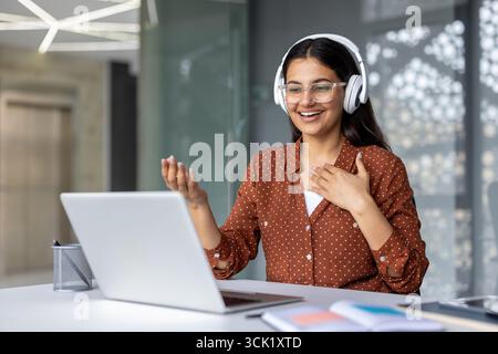 Young businesswoman wearing headphones, smiling while making a video call on her laptop, gesturing enthusiastically in a bright, modern office filled with elegant design elements Stock Photo