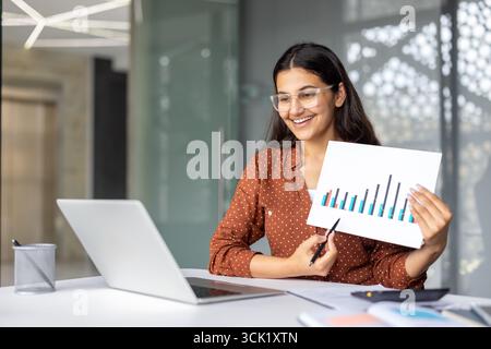 Young businesswoman pointing at colorful charts printed on paper while having video conference on laptop, sharing positive business results with her team Stock Photo