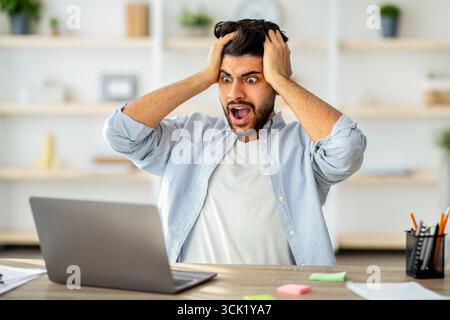 Deadline stress. Shocked arab man looking at laptop screen and touching head, suffering business problems Stock Photo