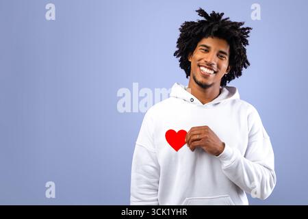 Expressing affection. Positive black teen guy holding small red paper heart on violet studio background Stock Photo