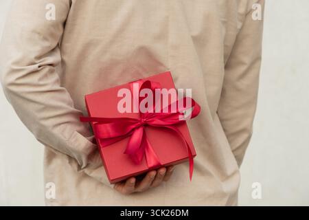 Man hiding red gift box tied with ribbon behind their back, wearing beige shirt for surprise girlfriend. Birthday present suggests surprise or special Stock Photo