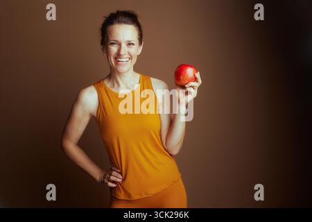 A vibrant woman in an orange tank top and leggings smiles widely, holding a red apple with her hand on her hip. She symbolizes healthy eating, fitness Stock Photo
