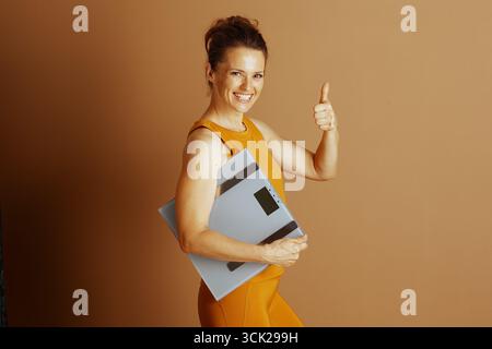 Smiling woman in activewear gives thumbs-up while holding digital scale, celebrating health and fitness achievements. Her positive gesture symbolizes Stock Photo