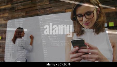 Working woman in white blouse writing on whiteboard in office, viewing code overlay on smartphone Stock Photo