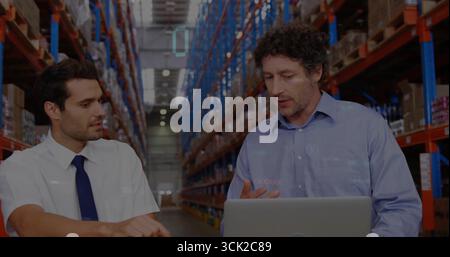 Reviewing stock data two men in business attire typing on laptop in warehouse aisle, with boxes Stock Photo