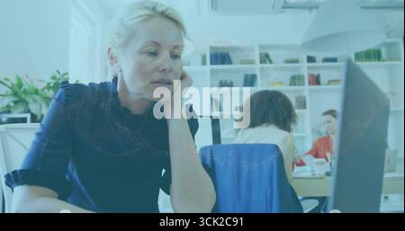 Leaning blonde-haired woman in navy blouse focusing on desktop monitor in open office, copy space Stock Photo