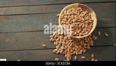 Spilling shelled almonds from round wooden bowl on rustic wooden plank tabletop, warm texture Stock Photo