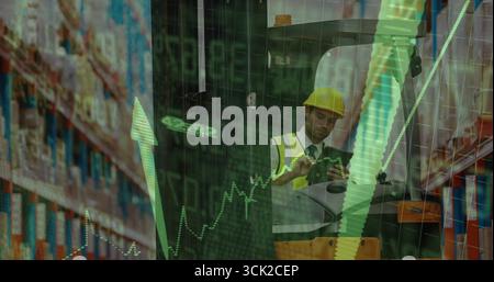 Manager wearing hard hat and vest using tablet in warehouse with forklift, racks and stock charts Stock Photo