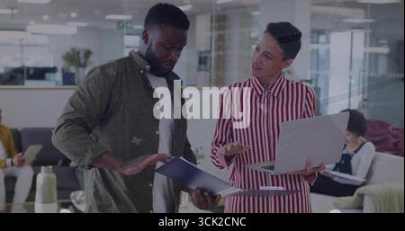 Reviewing blue document folder, two colleagues collaborating over silver laptop in office lounge Stock Photo