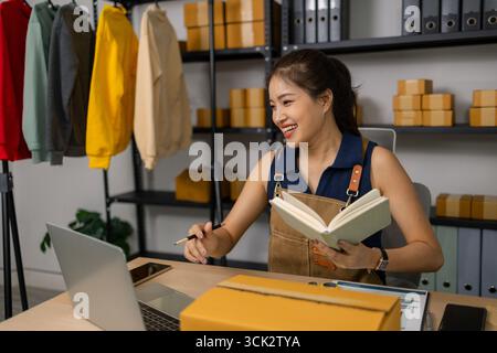 Young female business owner working happily at home, managing her online clothing store, checking orders and taking notes, using laptop to ensure succ Stock Photo