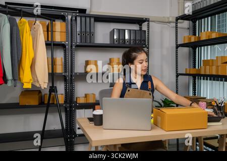 Young female entrepreneur working at home, managing her online clothing store business, checking orders from customers via laptop and preparing packag Stock Photo