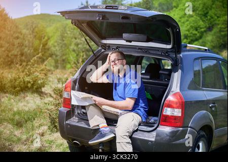 Man sits on open trunk of SUV in scenic, green landscape, holding map and scratching his head in confusion. Wearing blue shirt, beige pants, and colorful socks, he trying to figure out his location. Stock Photo