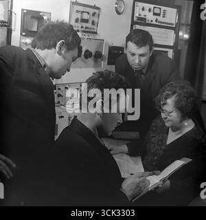 A group of Soviet engineers and technicians in a research and development lab or the central factory laboratory of the "Slavtyazhmash" plant (Sloviansk, Ukrainian SSR) discussing work results in February 1975. The archival photograph captures a team of specialists gathered around a table with a logbook, against a background of racks with control and measurement equipment. This scene reflects the spirit of the scientific and technical intelligentsia of the Era of Stagnation, emphasizing the importance of teamwork in solving production and research tasks. Stock Photo
