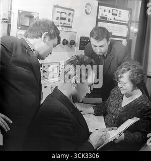 A group of Soviet engineers and technicians in a research and development lab or the central factory laboratory of the "Slavtyazhmash" plant (Sloviansk, Ukrainian SSR) discussing work results in February 1975. The archival photograph captures a team of specialists gathered around a table with a logbook, against a background of racks with control and measurement equipment. This scene reflects the spirit of the scientific and technical intelligentsia of the Era of Stagnation, emphasizing the importance of teamwork in solving production and research tasks Stock Photo