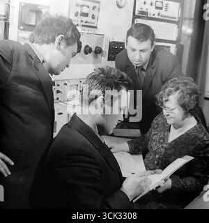 A group of Soviet engineers and technicians in a research and development lab or the central factory laboratory of the "Slavtyazhmash" plant (Sloviansk, Ukrainian SSR) discussing work results in February 1975. The archival photograph captures a team of specialists gathered around a table with a logbook, against a background of racks with control and measurement equipment. This scene reflects the spirit of the scientific and technical intelligentsia of the Era of Stagnation, emphasizing the importance of teamwork in solving production and research tasks Stock Photo