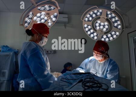 Two medical professionals perform a surgical procedure in a well-equipped operating room. They are dressed in sterile blue gowns and red caps, illumin Stock Photo